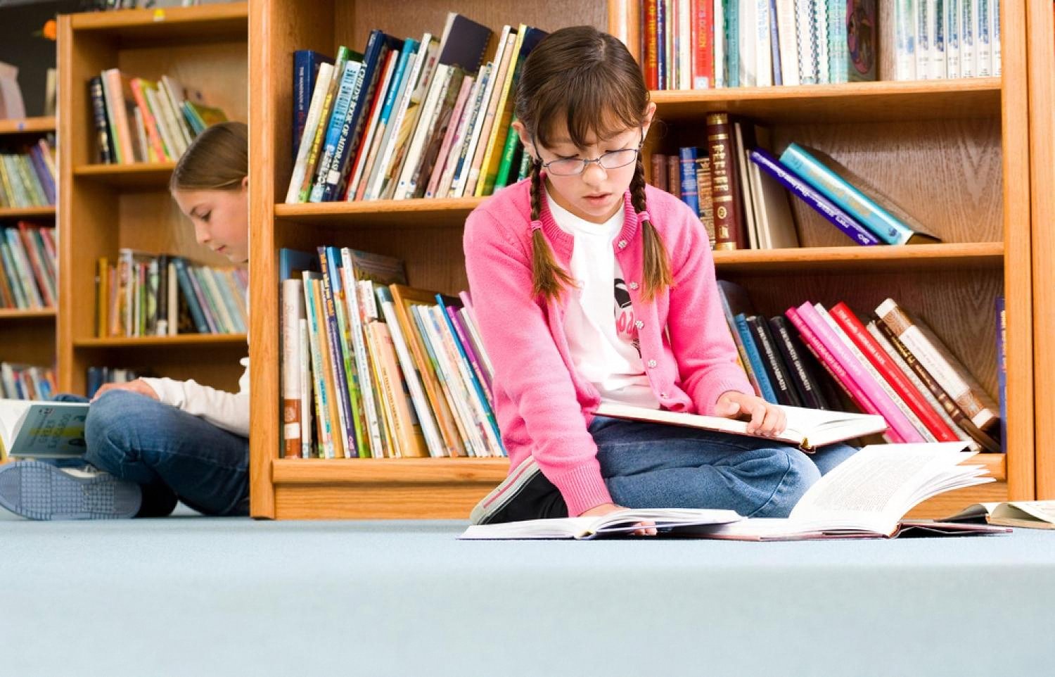 Two schoolgirls reading books on the floor of a school library